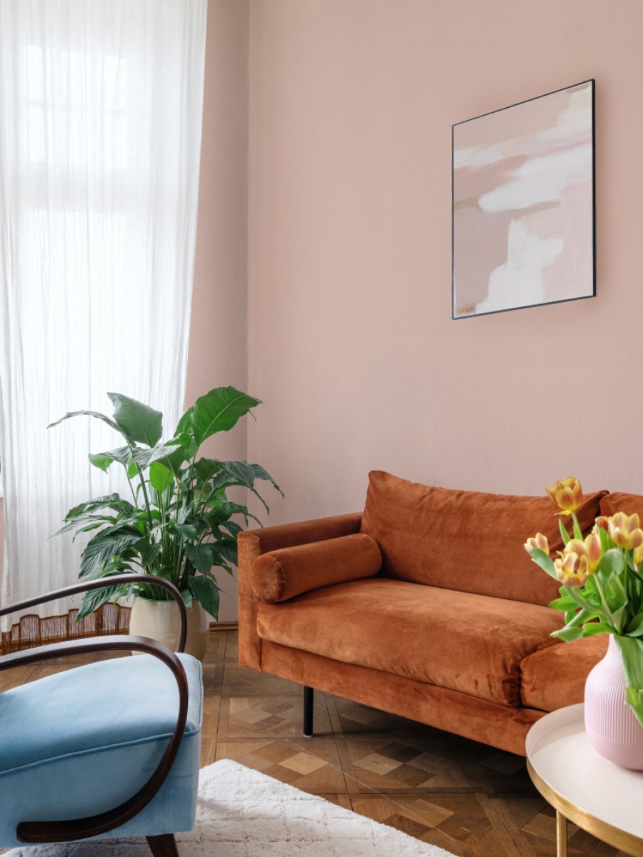 A living room with peach colored walls, a rust couch and blue armchair. There are beautiful house plants set around the room.