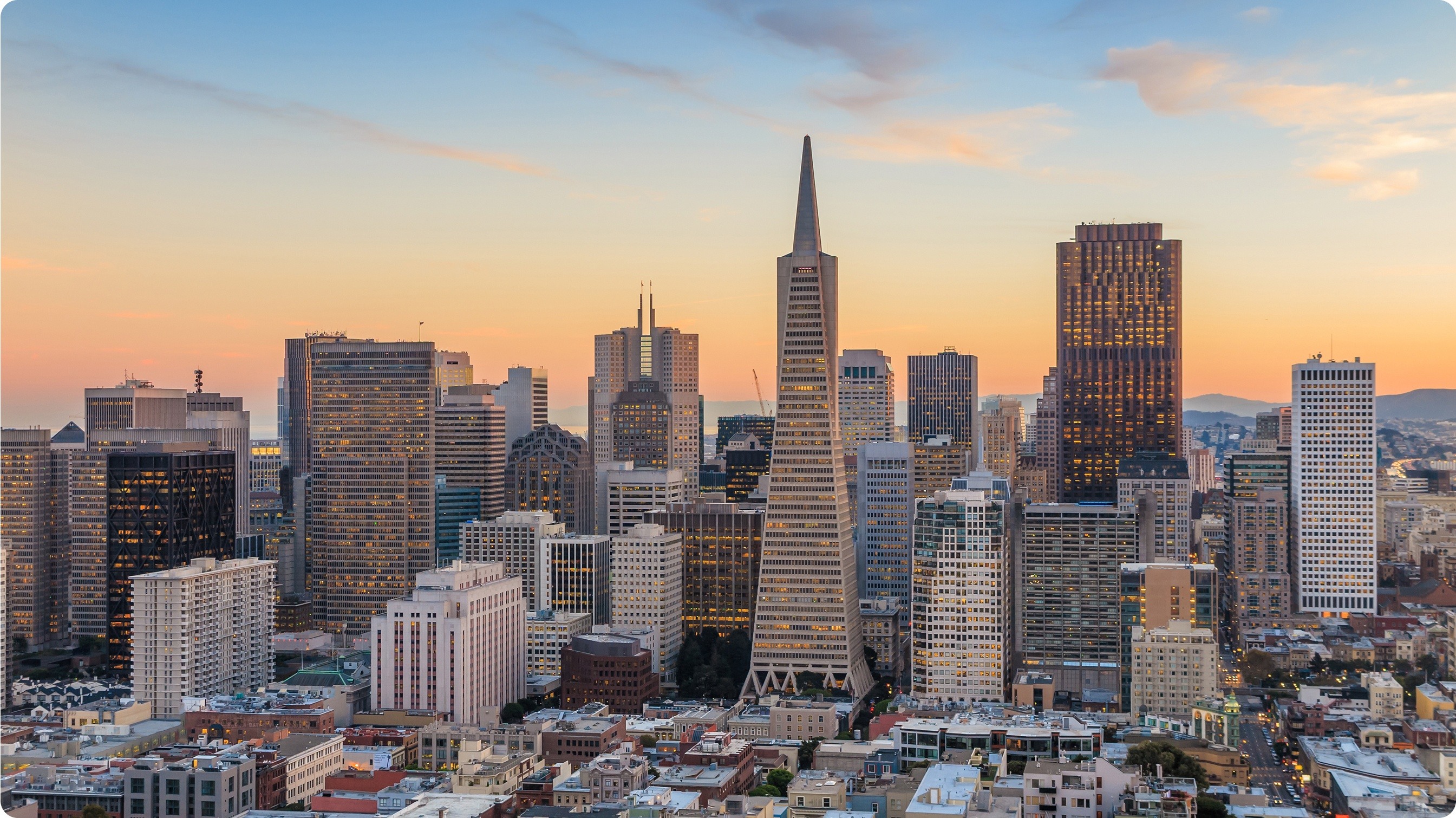San Francisco's Financial District photographed at dusk. Dozens of high-rise buildings, including the Transamerica Pyramid, sit in the mid-ground, with smaller, historic low-rise buildings in the foreground.