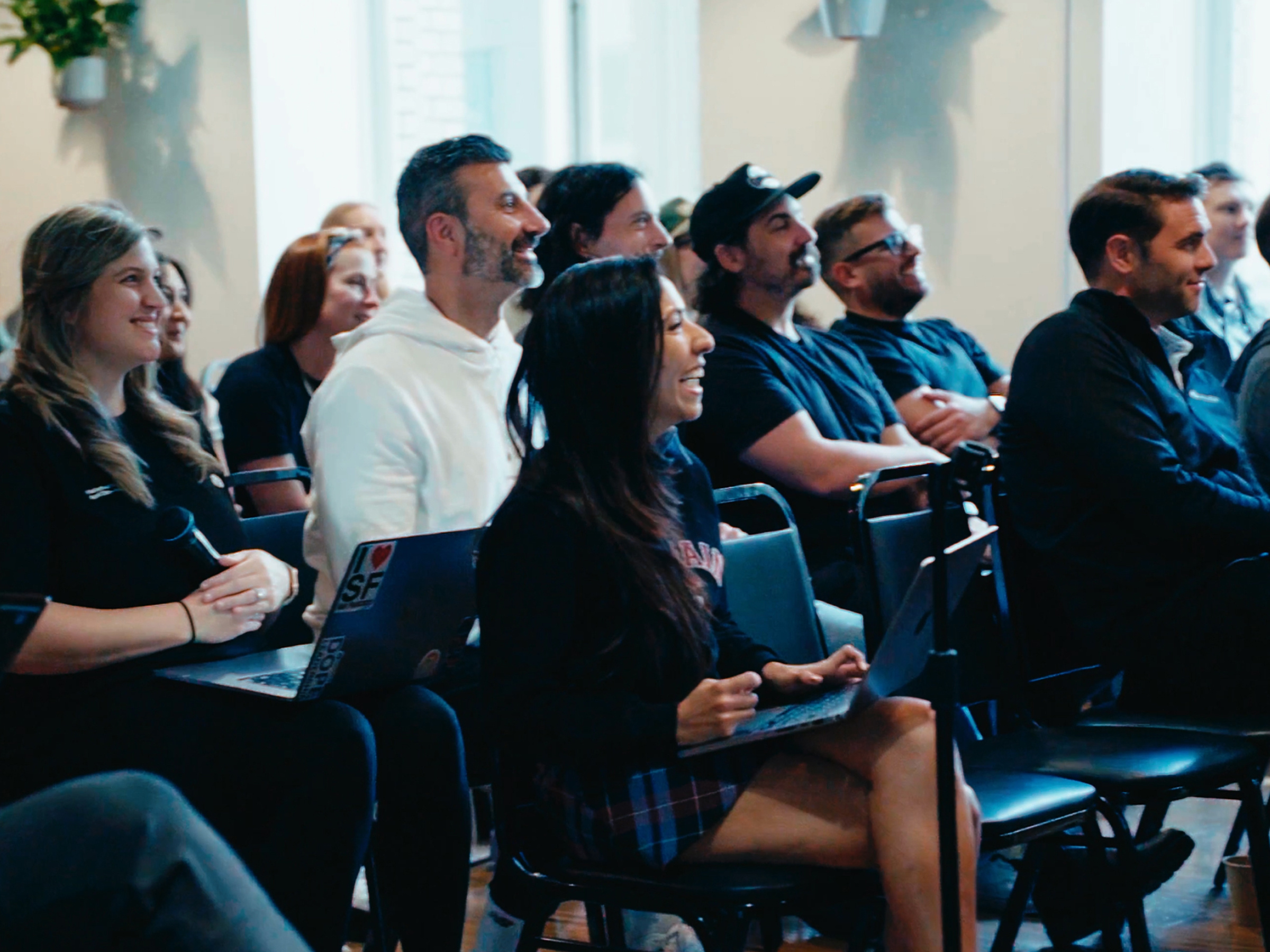A photo of a large group of Hover employees gathered in a communal area for a company-wide function. The people are all smiling and seated in chairs looking toward a speaker who is out of the frame.